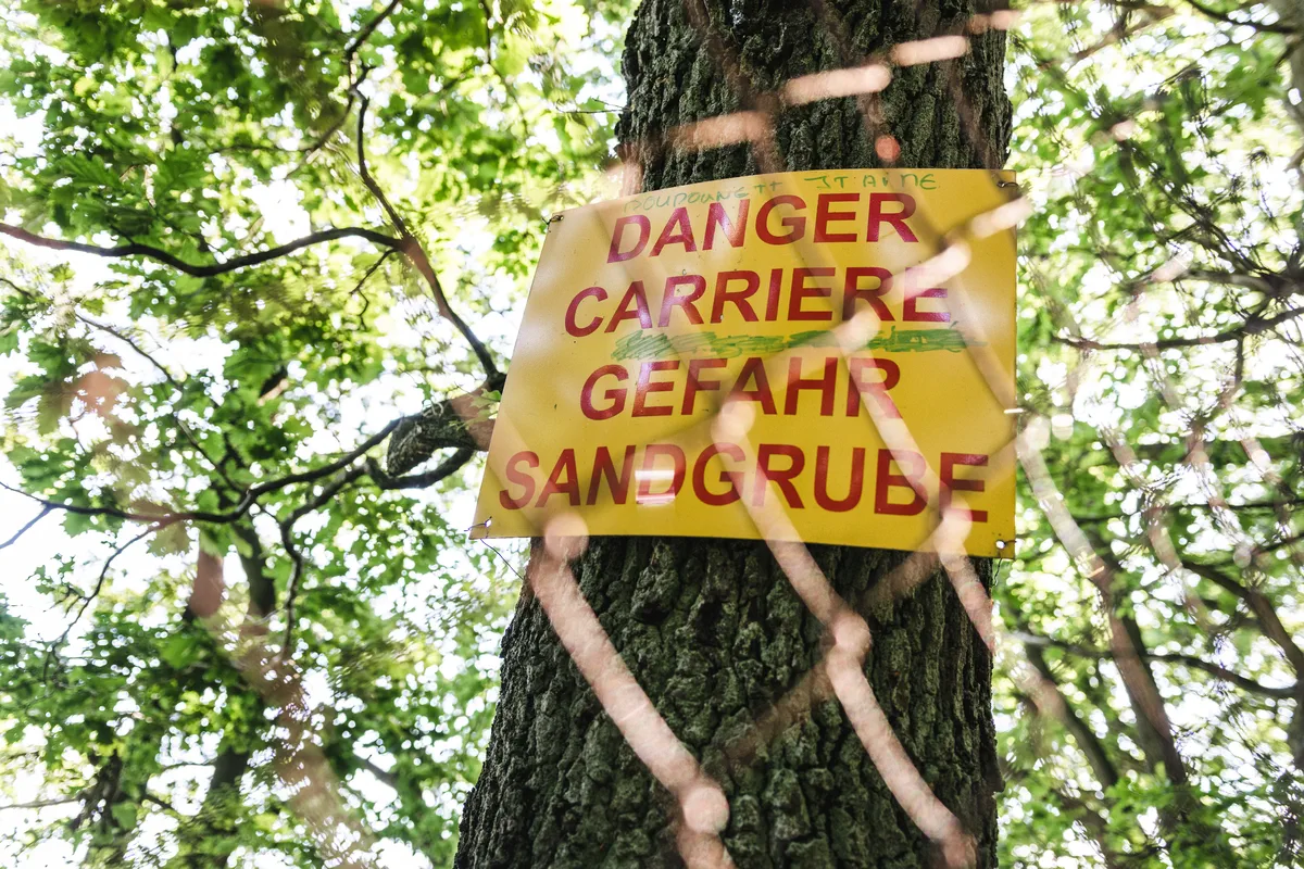Un panneau d'avertissement jaune attaché à un arbre indique "Danger, Carrière, Gefahr Sandgrube" en langues française et allemande. Le panneau est partiellement caché par un grillage. Des branches avec des feuilles vertes se trouvent en arrière-plan. Une série de panneaux identiques préviennent les promeneurs de la forêt du Warndt sur le danger de précipice dans la Carrière Barrois. Le sentier mène de Freyming-Merlebach à Karslbrunn.